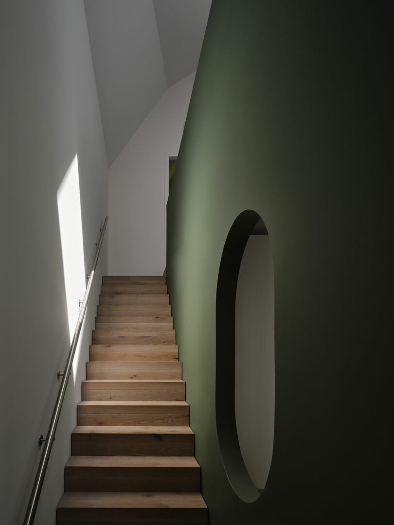 A wooden staircase with a metal handrail ascends beside a green wall with an oval opening, leading up to a brightly lit white wall and ceiling.