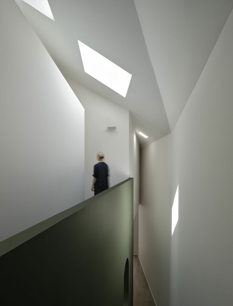 A person walks along a minimalist, white-walled corridor with geometric skylights and a green partition.