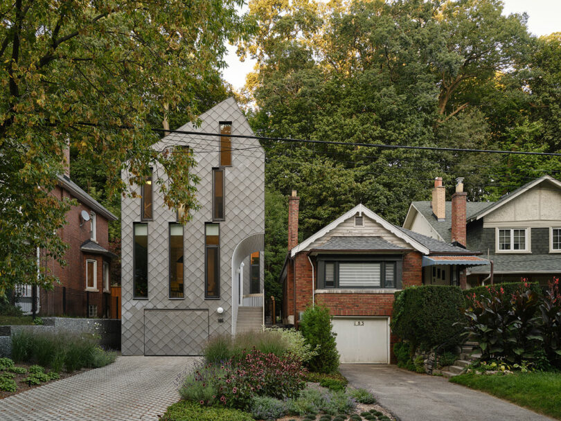 A modern, tall, narrow house with a gray, shingle-patterned exterior stands between two more traditional homes. There is a brick driveway in front, and lush greenery surrounds the house.