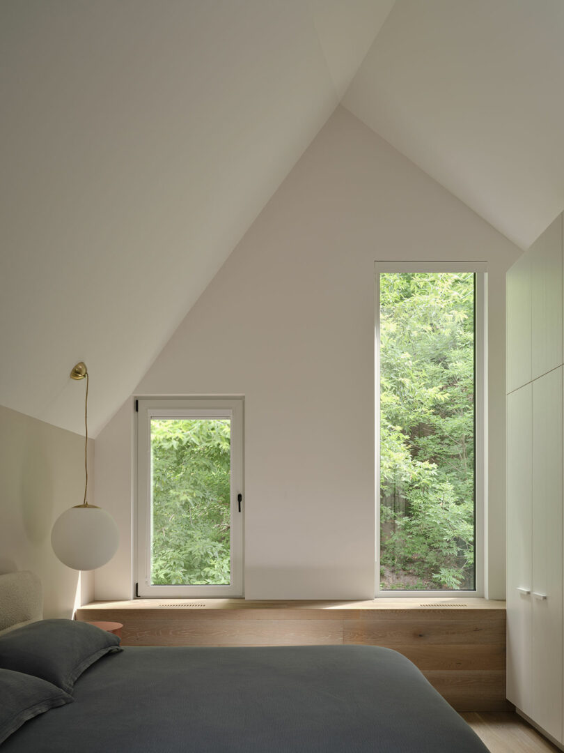 A minimalist bedroom with a slanted ceiling features two large windows with a view of green trees. The room is furnished with a bed, a pendant light, and built-in cabinetry.