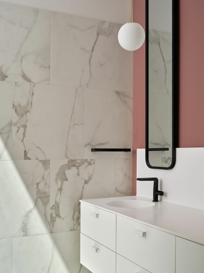 A modern bathroom features marble walls, a white sink with black fixtures, and a vertical mirror framed in black. A pendant light with a round white shade hangs above the sink.