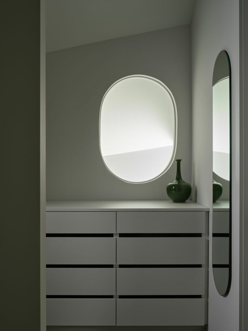 A minimalist room with a round window, a green vase on a white dresser, and an oval mirror on the wall.