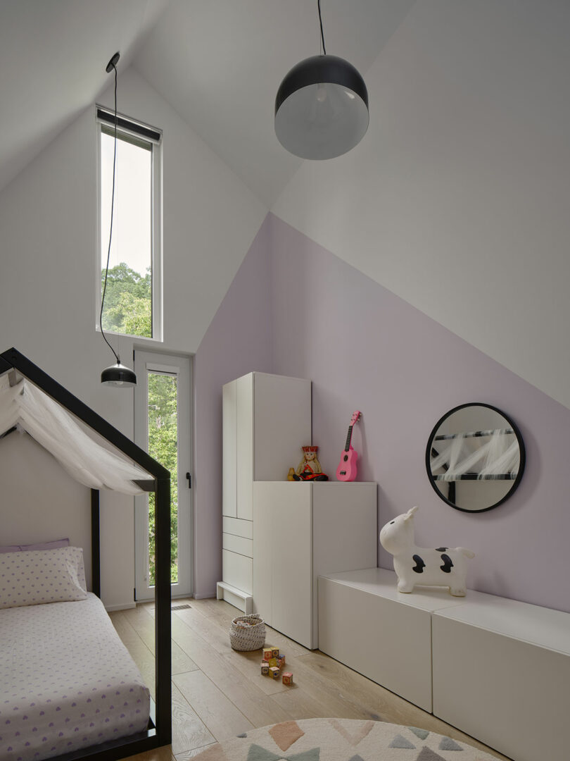 A children's bedroom with a bed, white cabinets, a pink guitar, a stuffed toy, and a round mirror. The room has a tall window and two black pendant lights. The walls are painted in shades of lavender and white.