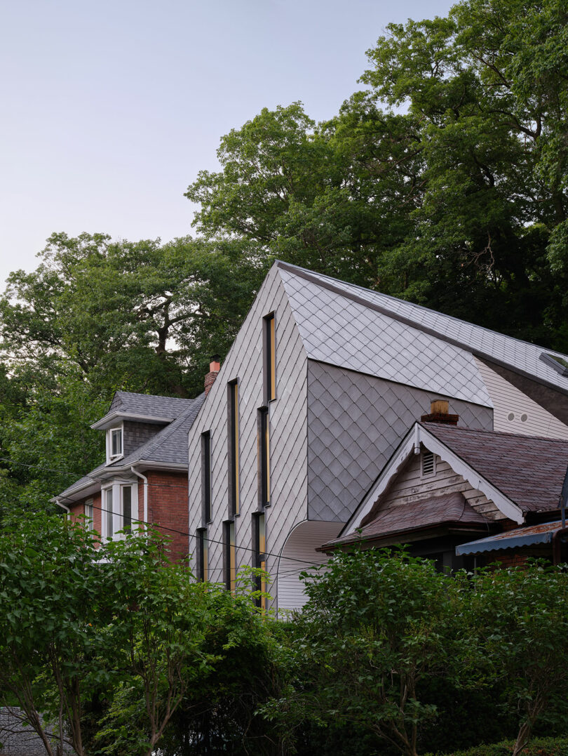 Modern house with a sloped metal roof and triangular windows positioned between two older, traditional homes; green trees and blue sky in the background.