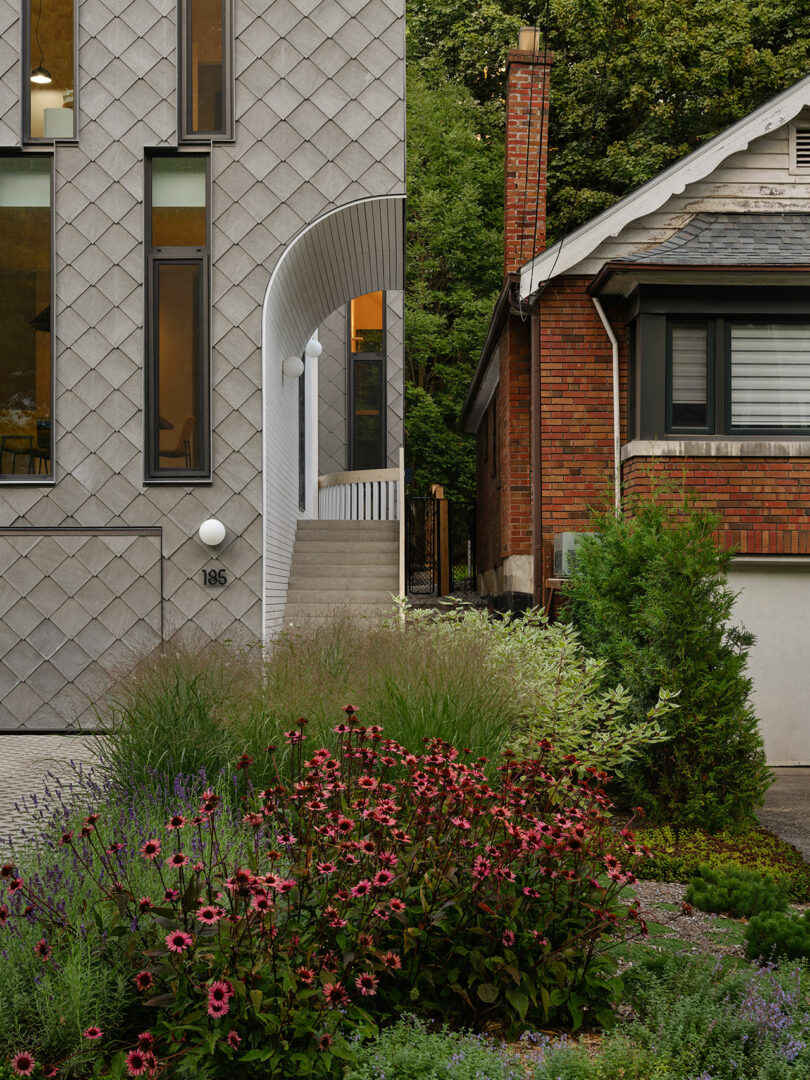 Modern gray house with large vertical windows and a staircase next to a traditional brick house. Both structures border a garden with various plants and pink flowers in front.