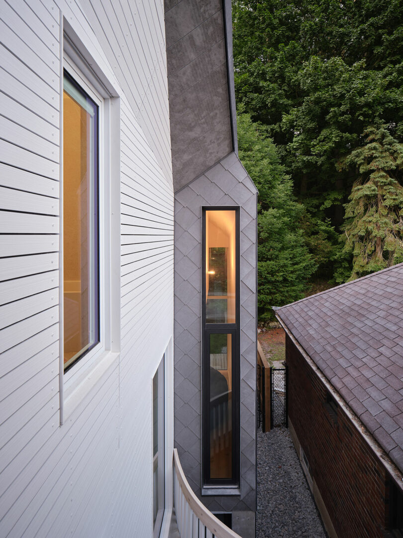 A narrow exterior corridor between a contemporary house with both white horizontal siding and gray shingles, and another building with a shingled roof, surrounded by dense green trees.