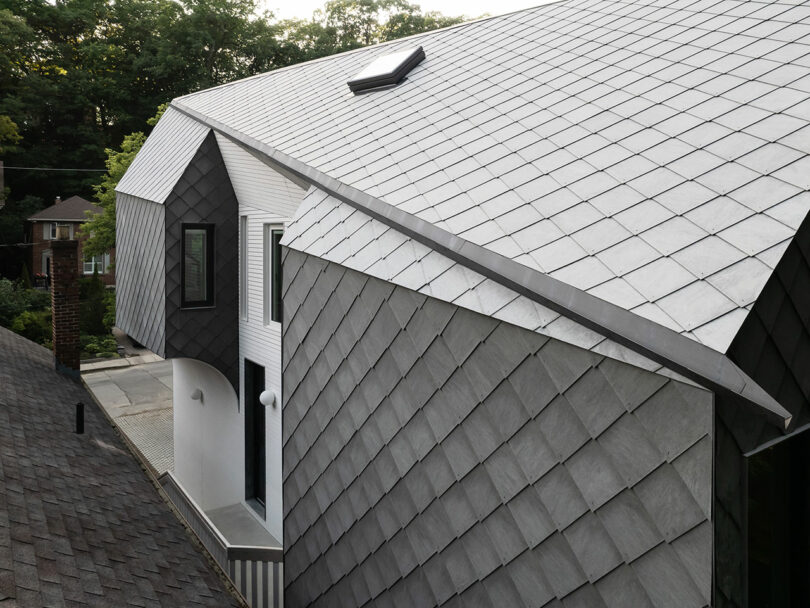 A modern house with a distinctive sloped roof covered in gray shingles. The exterior walls are also clad in matching shingles. There is a single window and skylight visible on the roof. Trees are in the background.