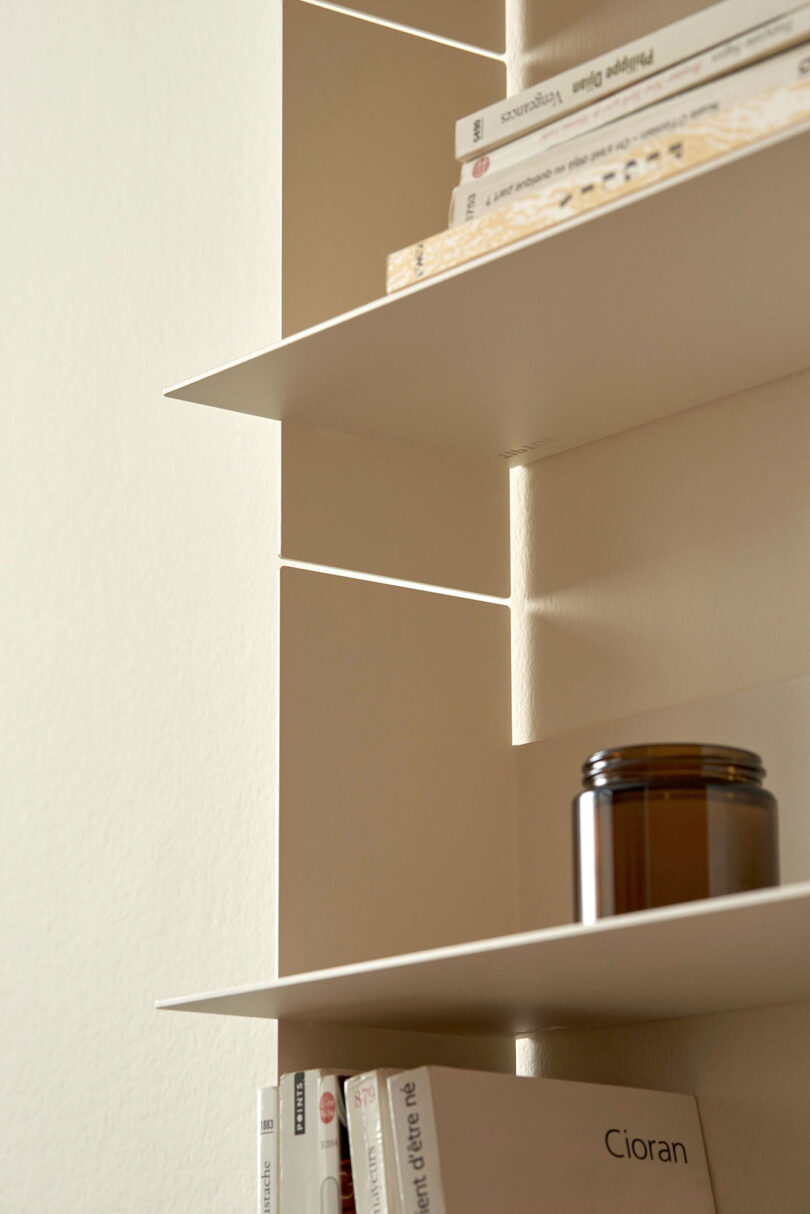 Close-up of a PLI Steel Wall Shelf by TIPTOE with several books and a brown jar placed on different shelves.