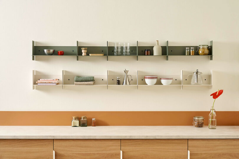 Minimalist kitchen with PLI Steel Wall Shelves by TIPTOE holding assorted kitchenware, including bowls, glassware, spices, and books. A countertop below displays jars and a single red flower in a vase.