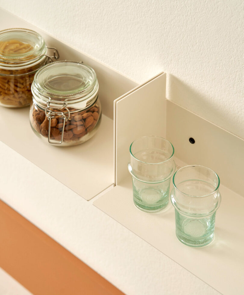 Two glass jars filled with food items and two empty glass tumblers are placed on a PLI Steel Wall Shelf by TIPTOE against a light-colored wall.