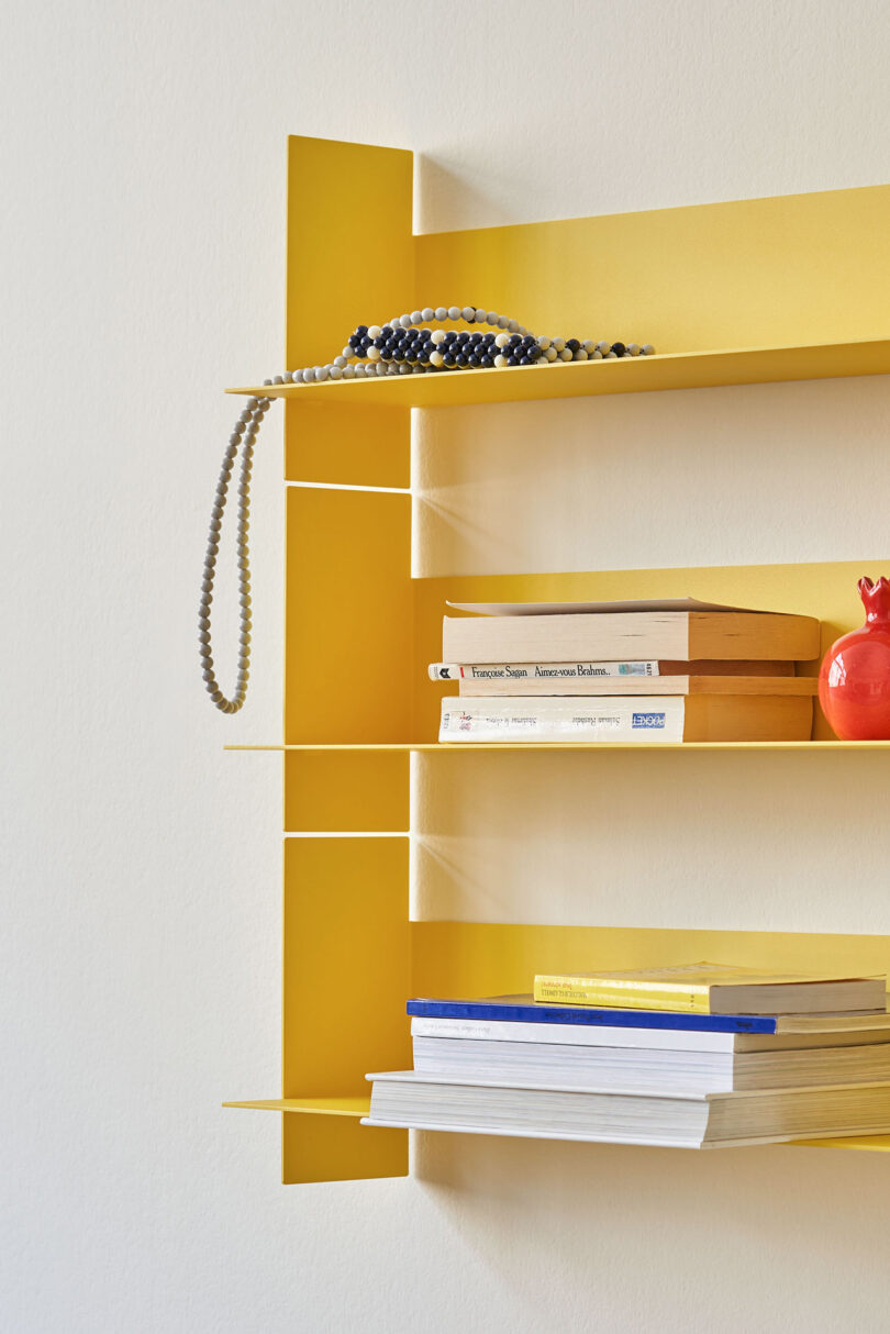 A yellow PLI Steel Wall Shelf by TIPTOE holds a decorative item, several books, and a necklace draped over one corner.