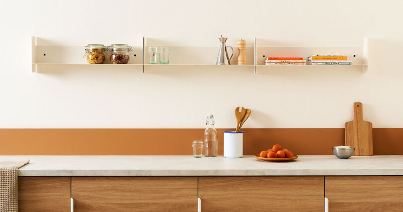 A minimalist kitchen with wooden cabinets, two white PLI Steel Shelves by TIPTOE holding jars and books, and a counter with kitchen utensils, a water bottle, two small jars, and a bowl of oranges.