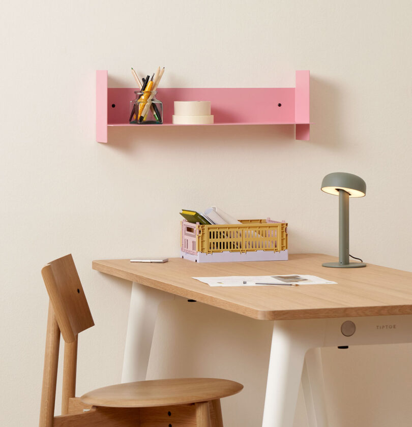 A minimalist workspace with a wooden desk, a matching chair, a small lamp, and stationery items. Above the desk, a PLI Steel Wall Shelf in Powder Pink holds a few objects, including a roll of tape and a small container.