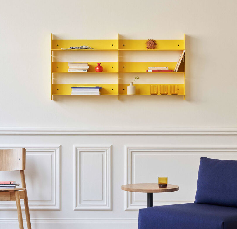 A yellow PLI Steel Wall Shelf by TIPTOE holds books, a red vase, a plant, and a sculpture. Below, a small wooden table with a glass sits next to a wooden chair and a blue couch against a cream wall.