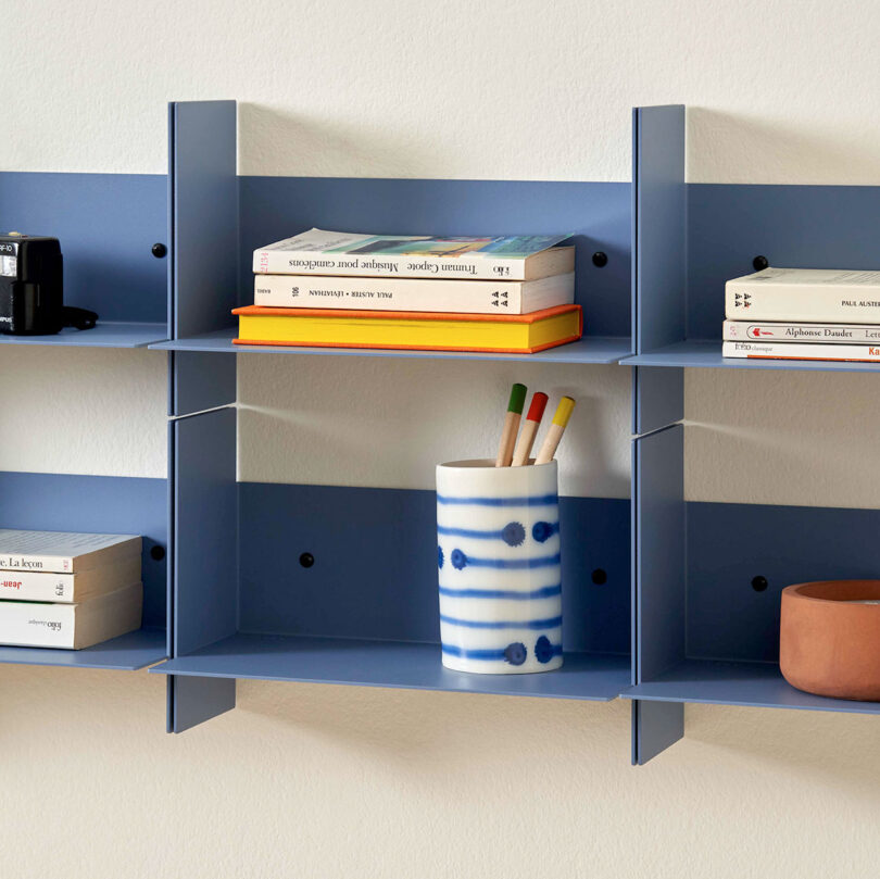 Blue PLI Steel Wall Shelves holding books, a camera, a blue and white pen holder with colored pencils, and a small orange cup.