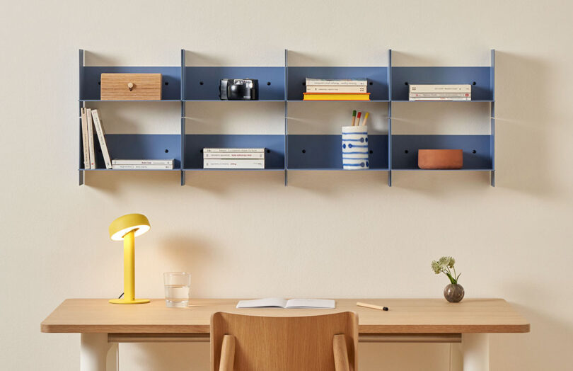 A wooden desk with a yellow lamp, a glass of water, an open notebook, and a small plant. Above, a blue PLI Steel Wall Shelf holds books, a camera, and various items. A wooden chair is tucked under the desk.