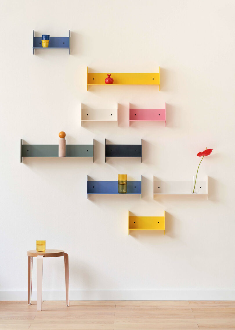 A minimalist interior with multicolored PLI Steel Wall Shelves holding various objects, and a single wooden stool with a yellow glass. One red flower is on the white shelf.