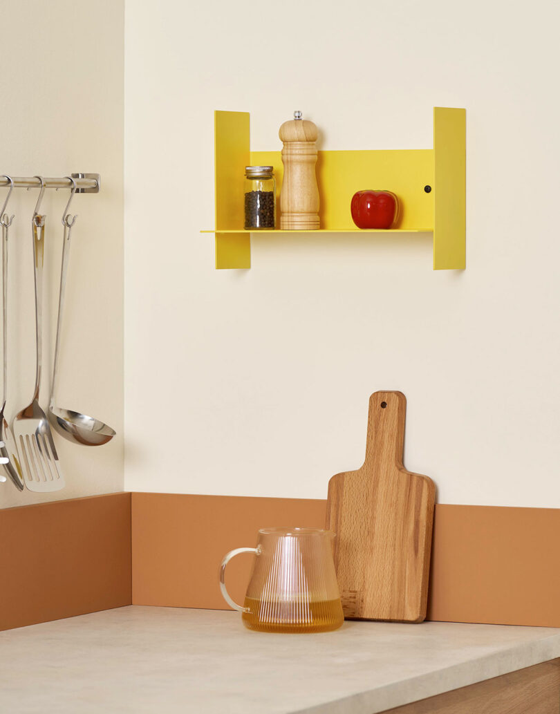 Kitchen scene with a yellow PLI Steel Wall Shelf holding a pepper grinder, small jar, and a red tomato. Utensils hang on the left. A cutting board and a glass pitcher are on the counter below.