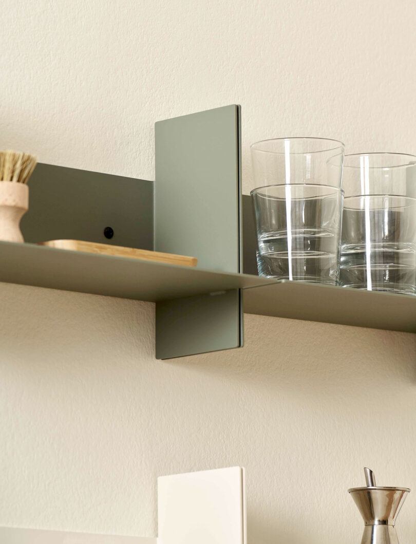 A green PLI Steel Wall Shelf by TIPTOE holds two clear glass tumblers, a scrubbing brush, and a square wooden item.