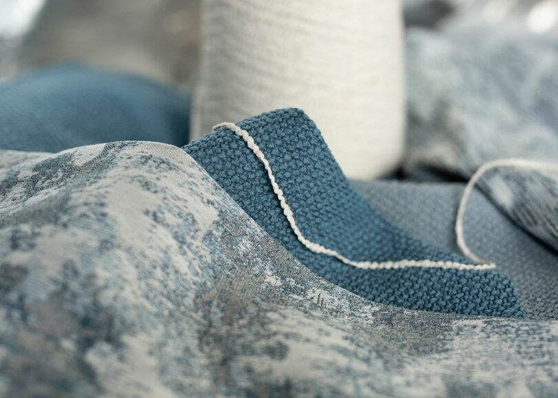 Close-up of blue and gray textured fabric with white stitching, draped on a surface. A roll of white yarn is visible in the background.