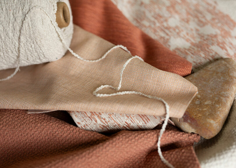 A close-up of various textured fabrics in earthy tones with a spool of thread and a stone on top.