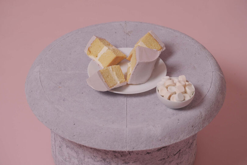 Three slices of layered cake on a white plate, next to a small bowl of white marshmallows, on the stone-textured surface of the Pastille Table by Emeline Ong against a pink background.