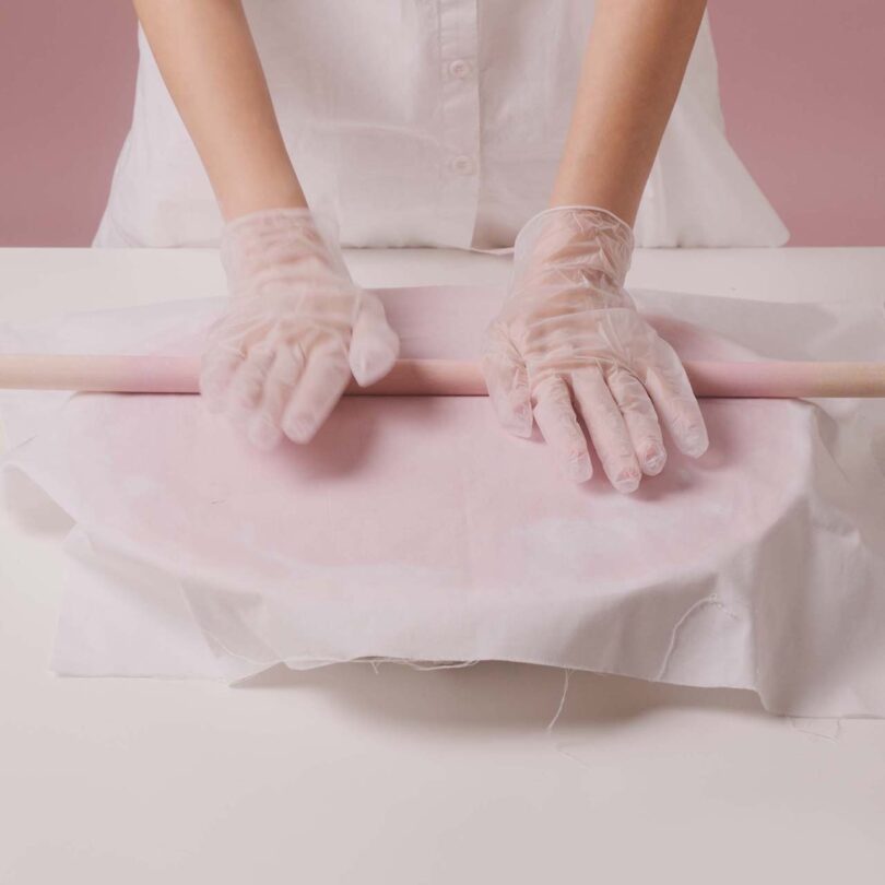 Emeline Ong wearing white gloves uses a rolling pin to flatten pink paper composite on a white surface.