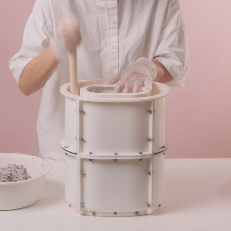 Emeline Ong, wearing gloves, mixes materials in a white cylindrical mold with multiple metal screws on a table, with a pink background. A bowl with a gray substance is placed nearby.