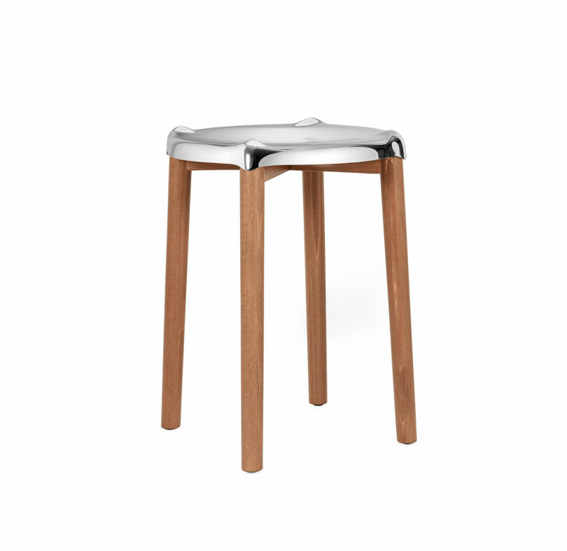 The Poele Stool with a polished metal seat and four wooden legs standing against a white background.