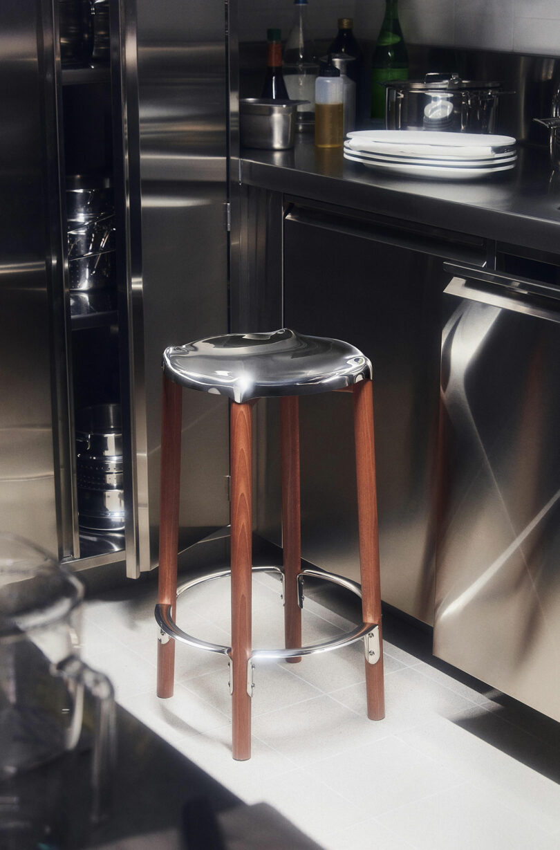 The Poele High Stool by Philip Starck and Alessi with a polished metallic seat stands on a tiled floor in a modern kitchen. The stool is surrounded by stainless steel cabinets and appliances.