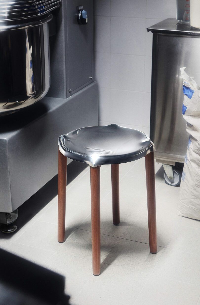 The Poele Stool by Philip Starck and Alessi with a shiny, black seat and wooden legs is positioned on a light grey floor in an indoor setting, surrounded by industrial equipment and a bag.