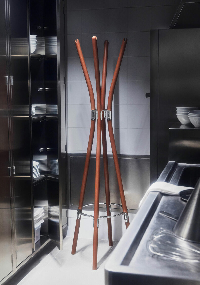 The Poele Coat Rack stands in a stainless steel industrial kitchen near a shelf with stacked plates and bowls.