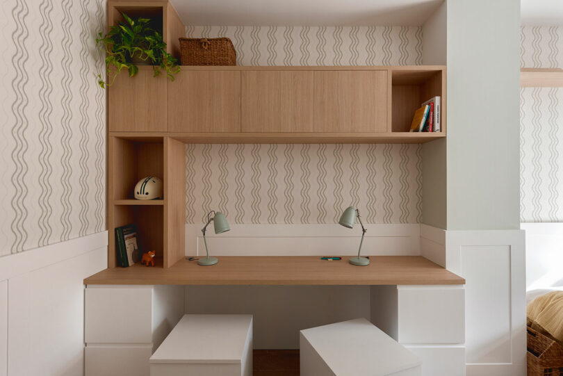 A modern study area featuring a wood and white desk with twin lamps, open and closed storage shelves, a potted plant, and a few books against a wavy patterned wall.