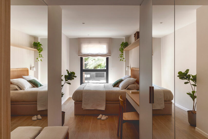 A modern bedroom with a double bed, two plants, a desk, and a large window. The room features light-colored decor, a bench at the foot of the bed, and mirrored closets.