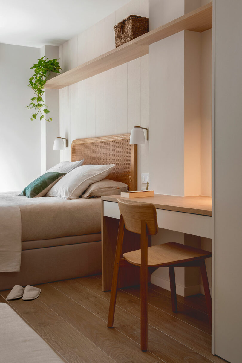 A cozy bedroom features a bed with beige linens and pillows, a wooden desk with a chair, wall-mounted lamps, a shelving unit with a wicker basket, and a hanging plant.