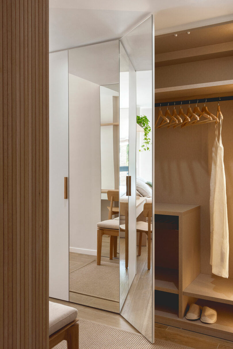 A minimalist bedroom with a mirrored wardrobe door, wooden chair, hanging beige dress, and open shelves.