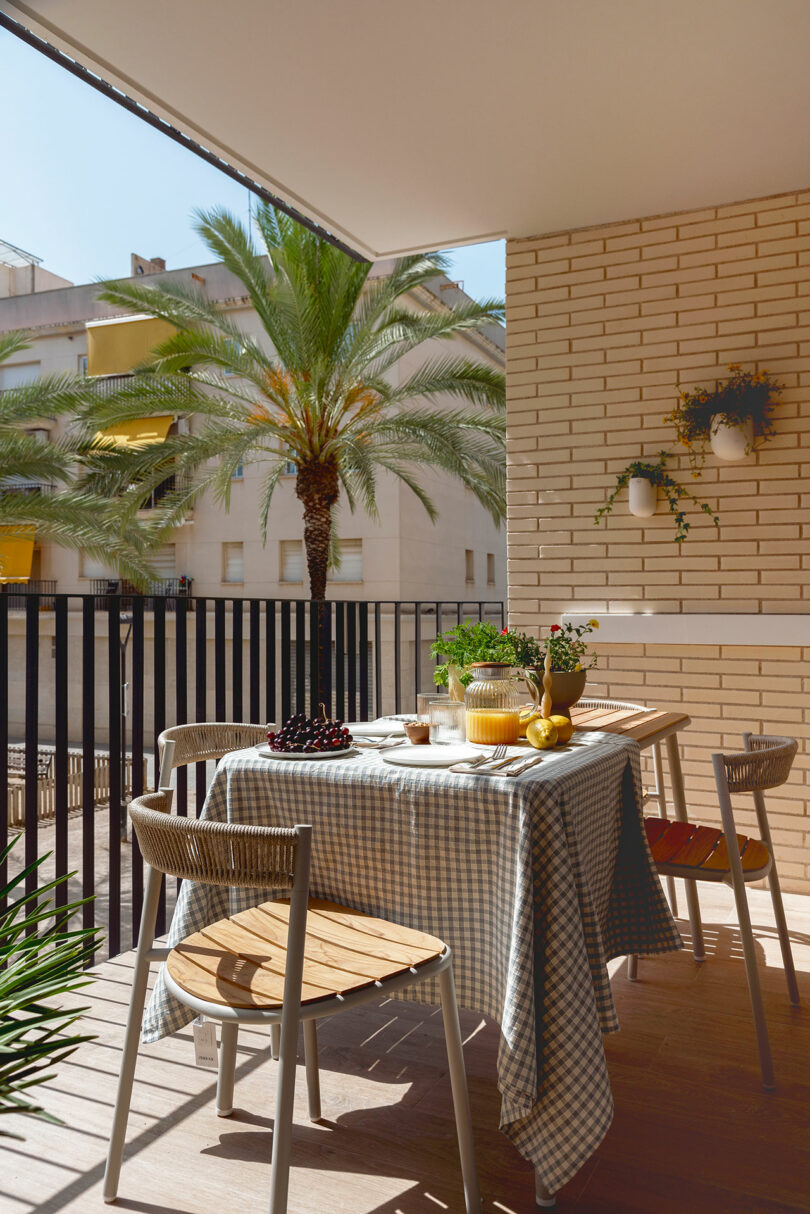 A sunlit balcony with a table covered by a checkered tablecloth, adorned with potted plants and flowers. Two chairs are positioned beside the table, and a palm tree is visible in the background.