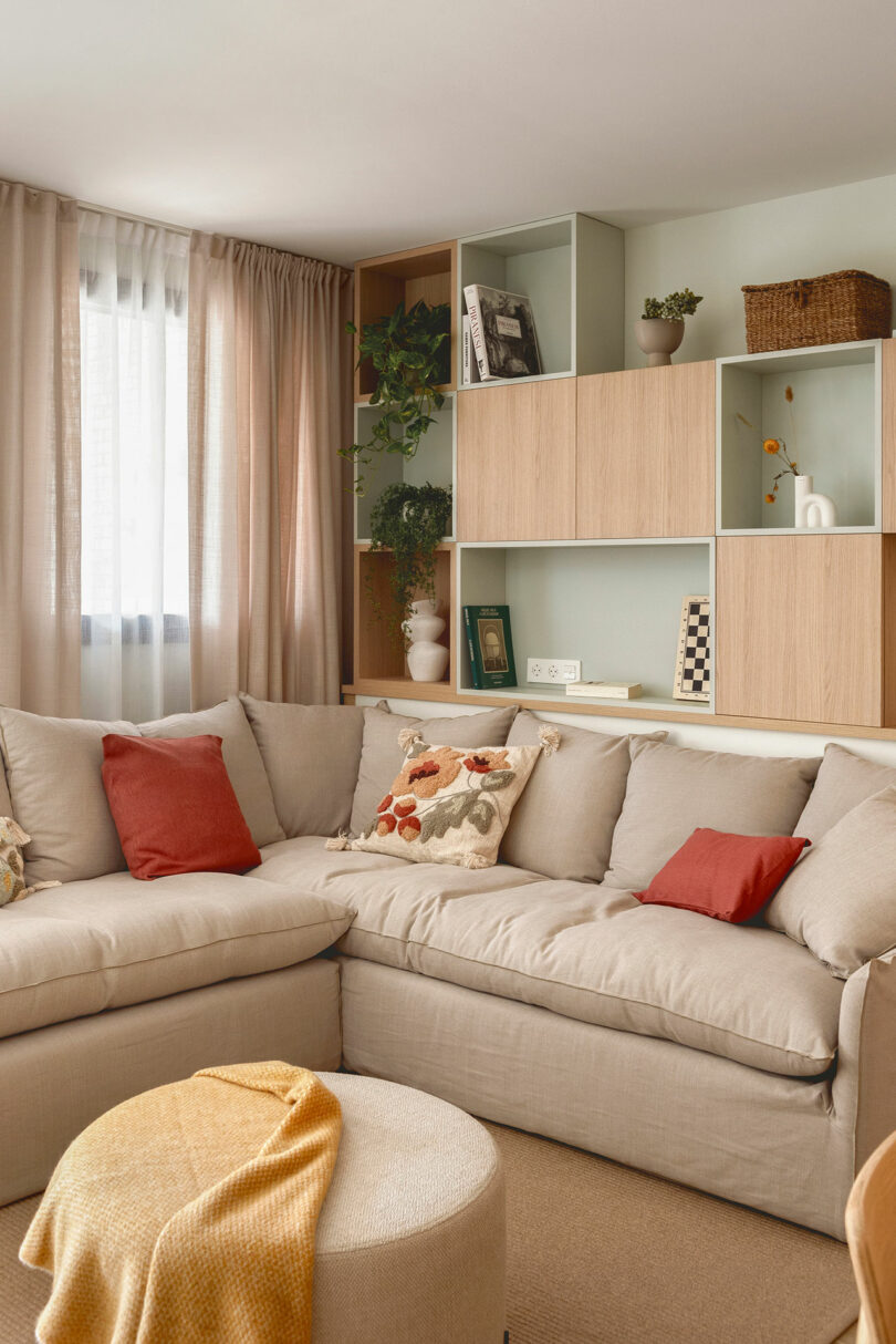 A cozy living room with a beige sectional sofa adorned with red and floral pillows, a round beige ottoman with a yellow throw blanket, and a wooden shelving unit with various decorations and plants.