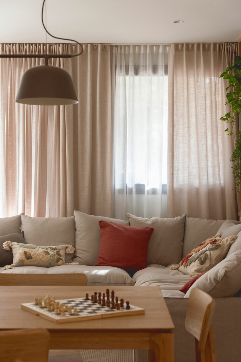 A cozy living room with a light gray sectional sofa featuring colorful pillows, a wooden table with a chessboard, and a large window with sheer curtains.