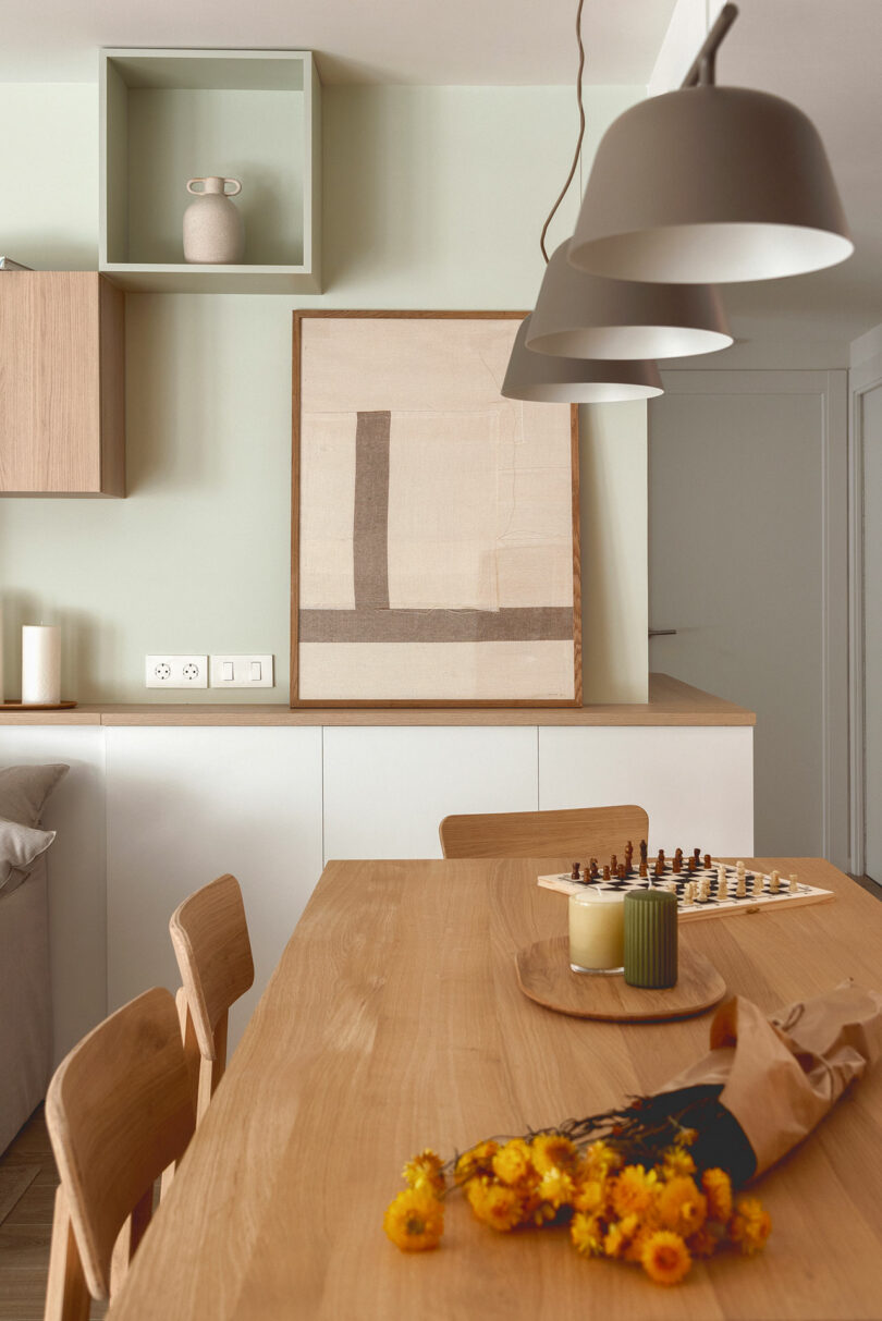 A modern dining area with a wooden table, chairs, a chessboard, a vase with yellow flowers, and a minimalist wall art. Pendant lights hang above the table, and cabinetry is in the background.