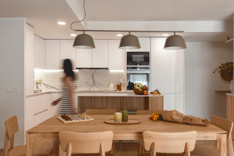 Modern kitchen and dining area with light wooden furniture, marble backsplash, and pendant lights. A person in motion, blurred, moves near the counter which is decorated with fruits and vegetables.