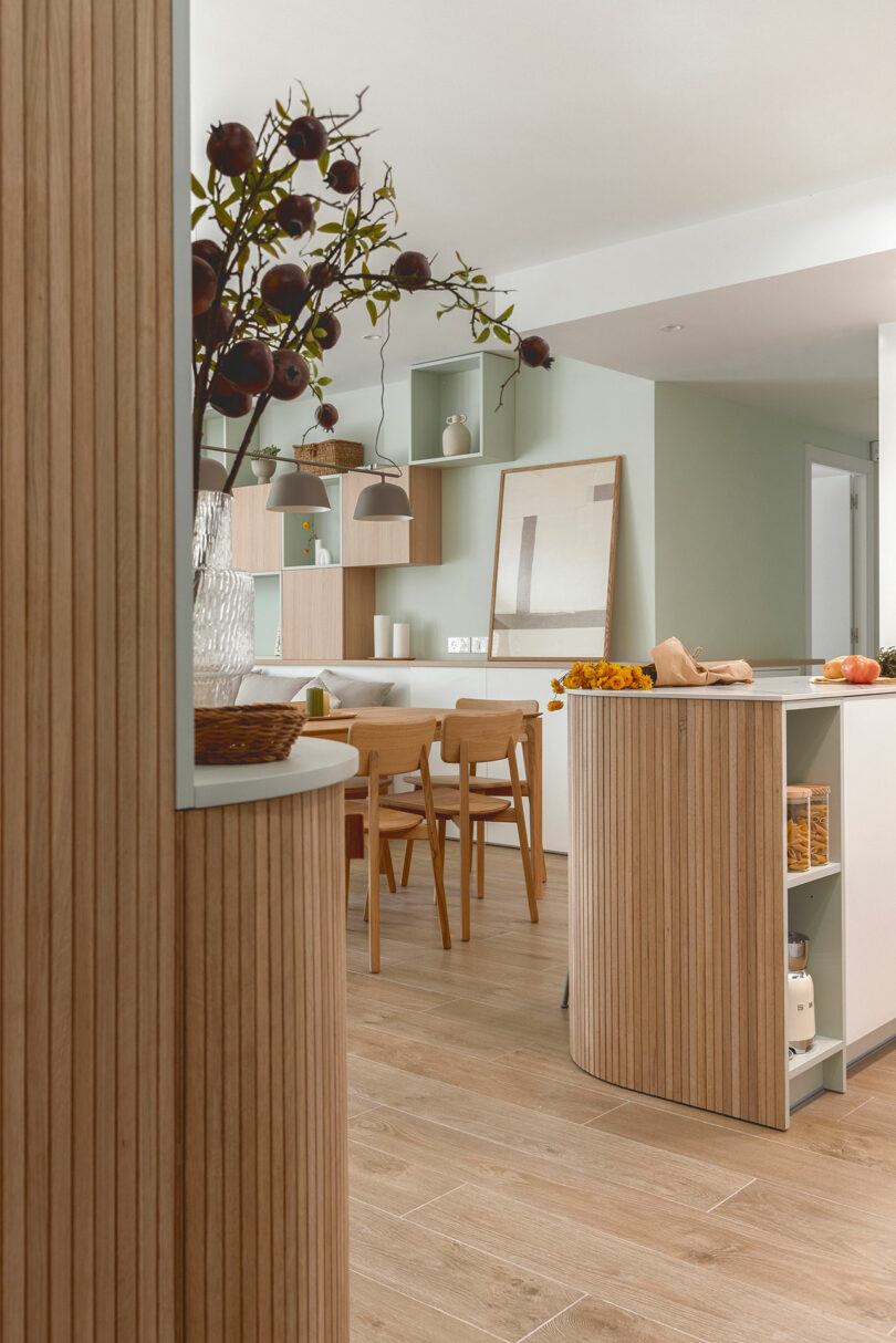 A modern kitchen and dining area with light wood furnishings, green walls, and minimalist decor. There are chairs around a table, shelves with items, and a vase with branches on the wooden counter.