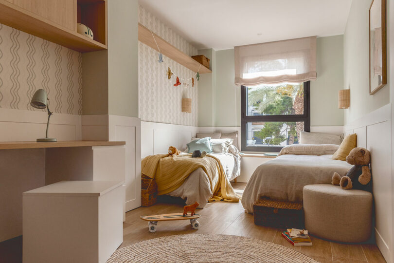 A cozy children's bedroom with two beds, a skateboard on the floor, shelves with decorations, a desk with a lamp, a cushioned stool, and a window letting in natural light.