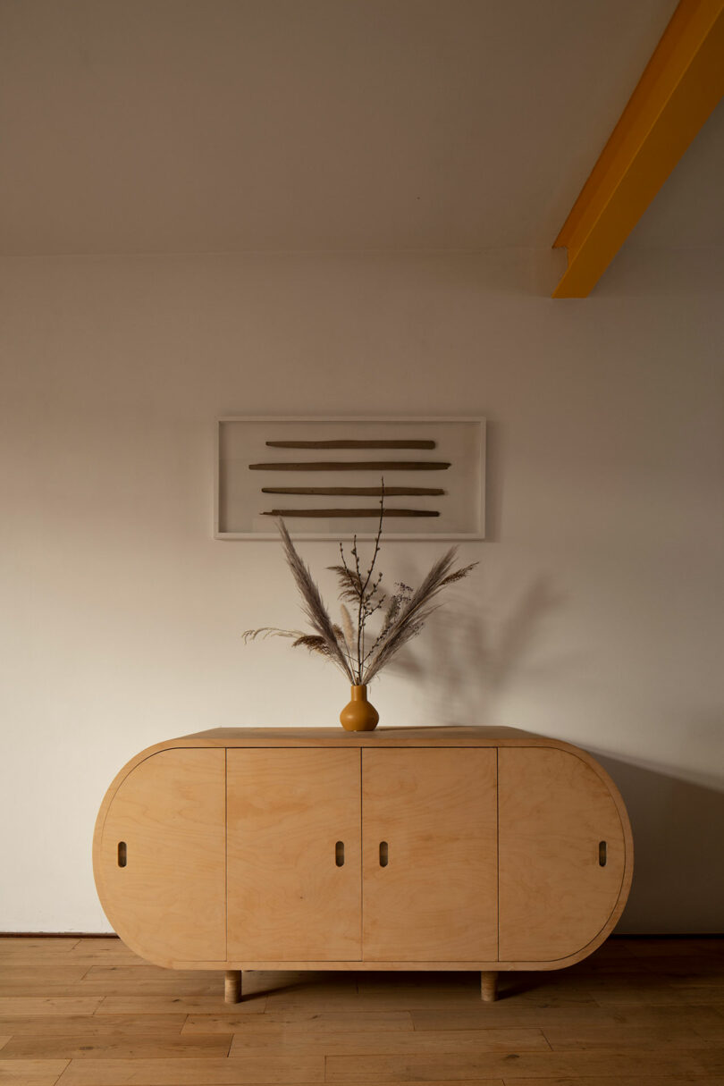 A wooden sideboard with round edges is topped with a ceramic vase holding dried plants. Above it, there is a framed piece of minimalist art with horizontal lines hanging on a white wall.