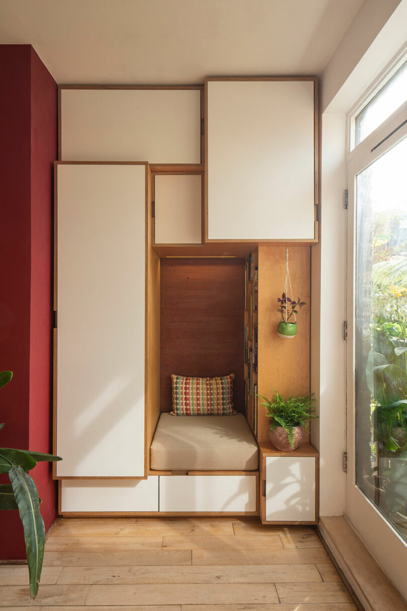 A small, minimalist reading nook with a cushion, surrounded by white cabinets and a red accent wall, next to a sunlit window. Houseplants are arranged on a low shelf and hanging from the wall.