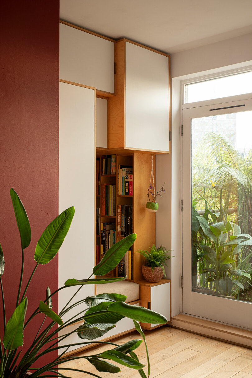 A modern interior space with built-in wooden shelves filled with books and plants, adjacent to a glass door and surrounded by various indoor plants.