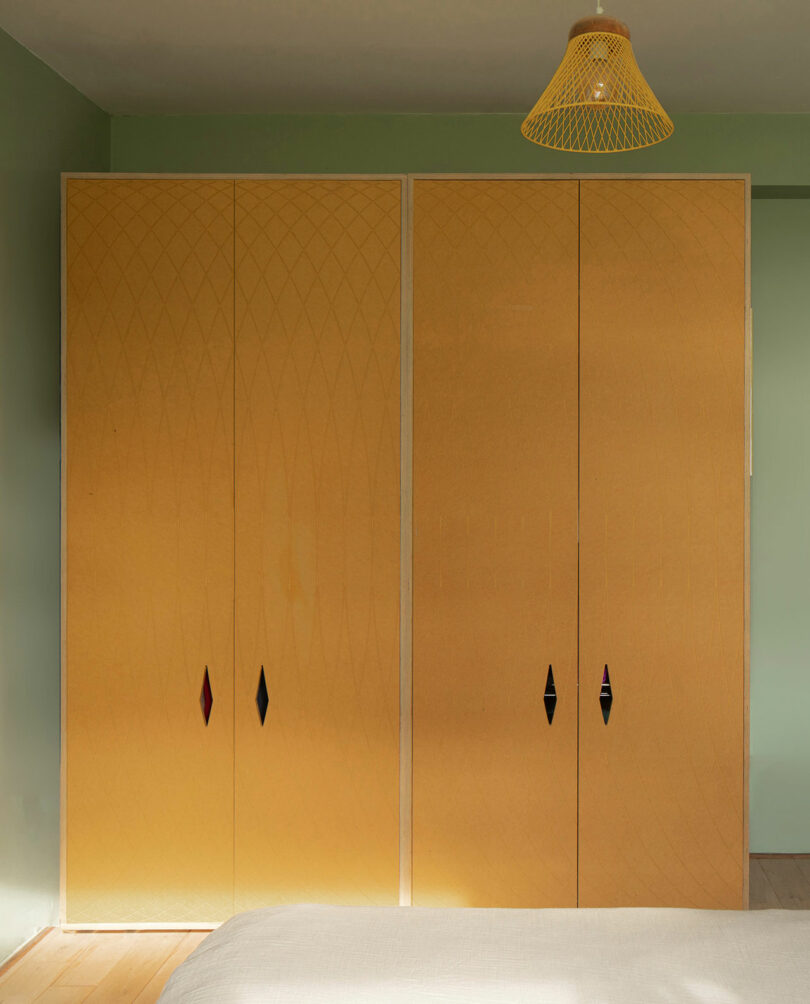 A minimalist bedroom features a wooden wardrobe with subtle diamond-patterned doors, black and red diamond-shaped handles, and a yellow woven pendant light hanging from the ceiling.