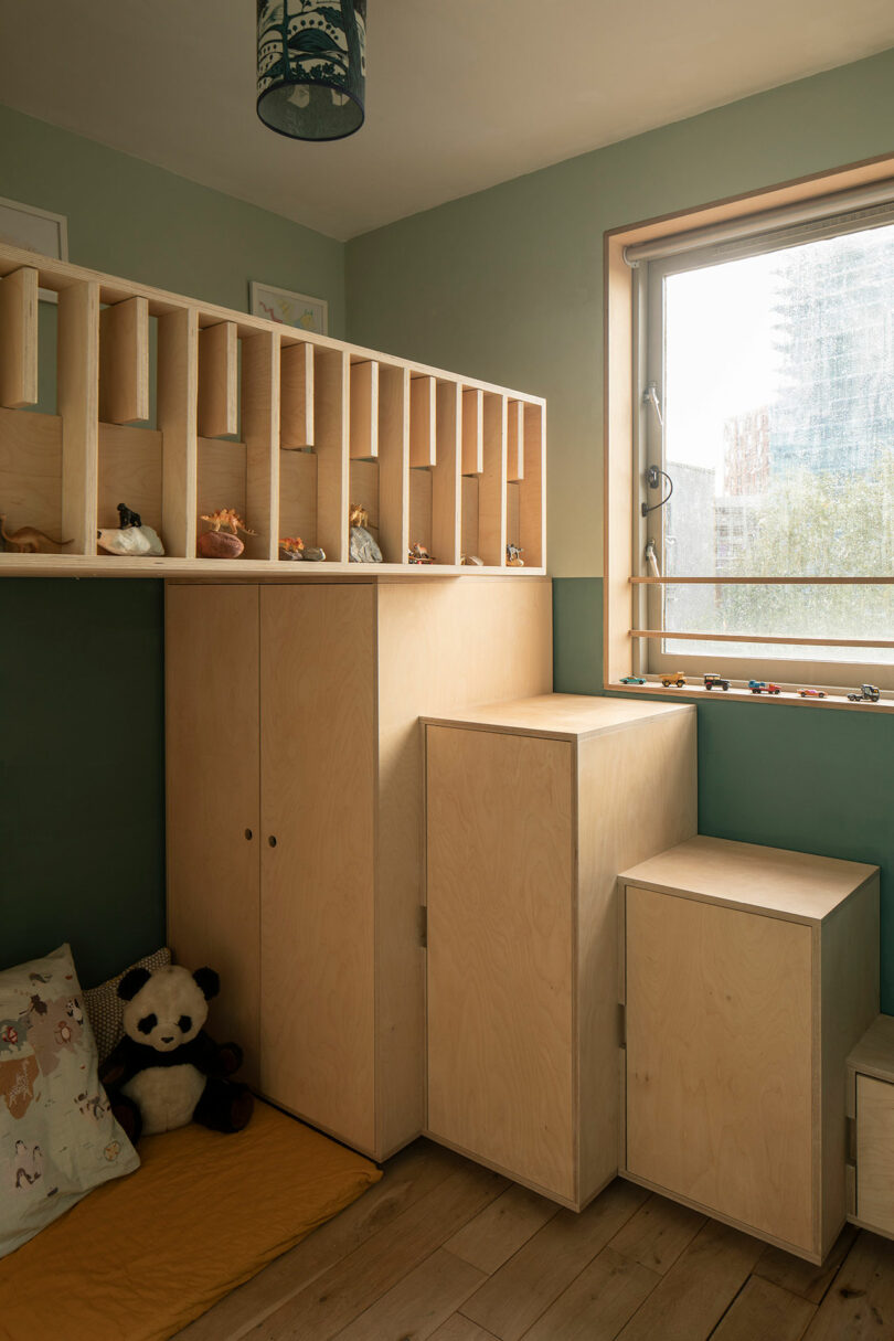 A small children's room with wooden shelves displaying toys, a cushioned area with stuffed animals, and steps leading up to a window. The walls are painted green and light filters through the window.