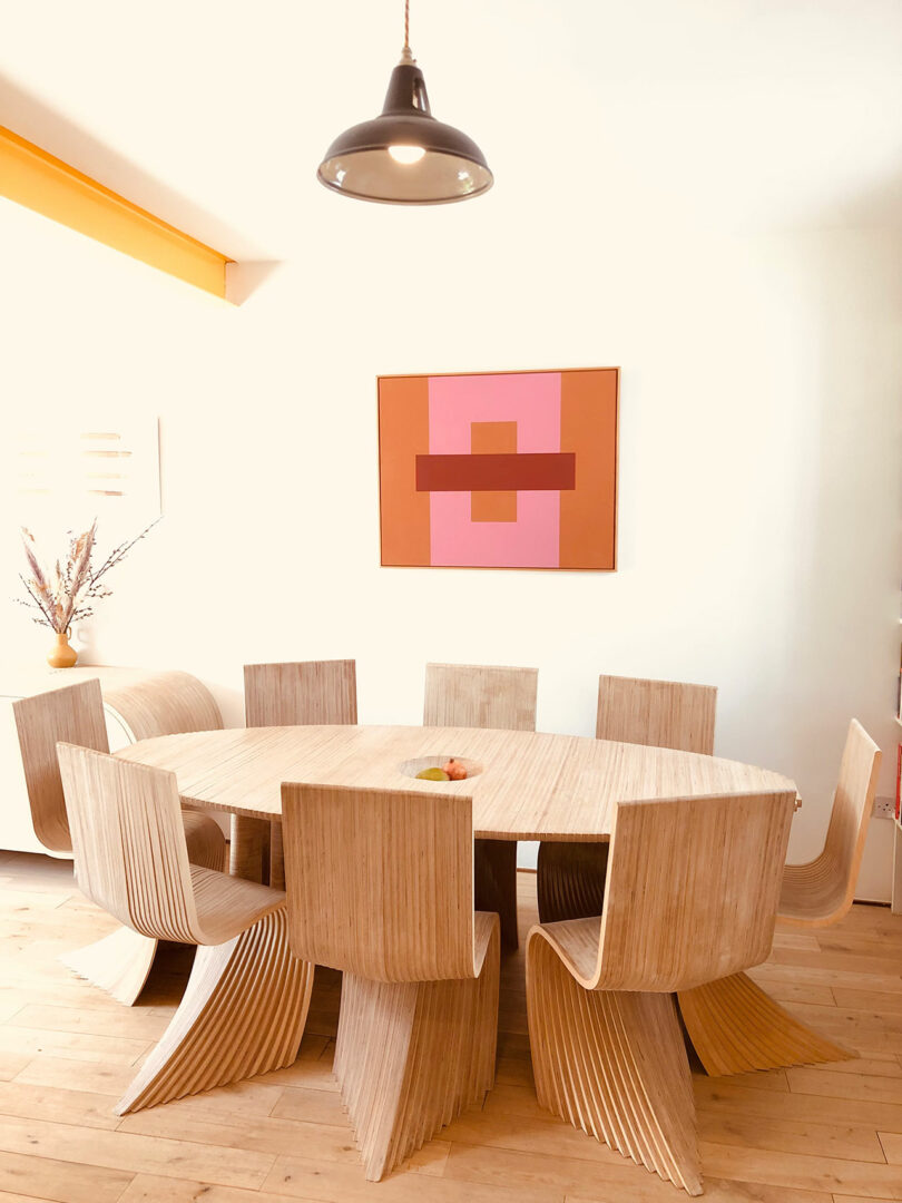 A modern dining room with a round wooden table and eight curved wooden chairs. A geometric painting is on the wall, and a bowl with fruit is on the table. A pendant light hangs above.