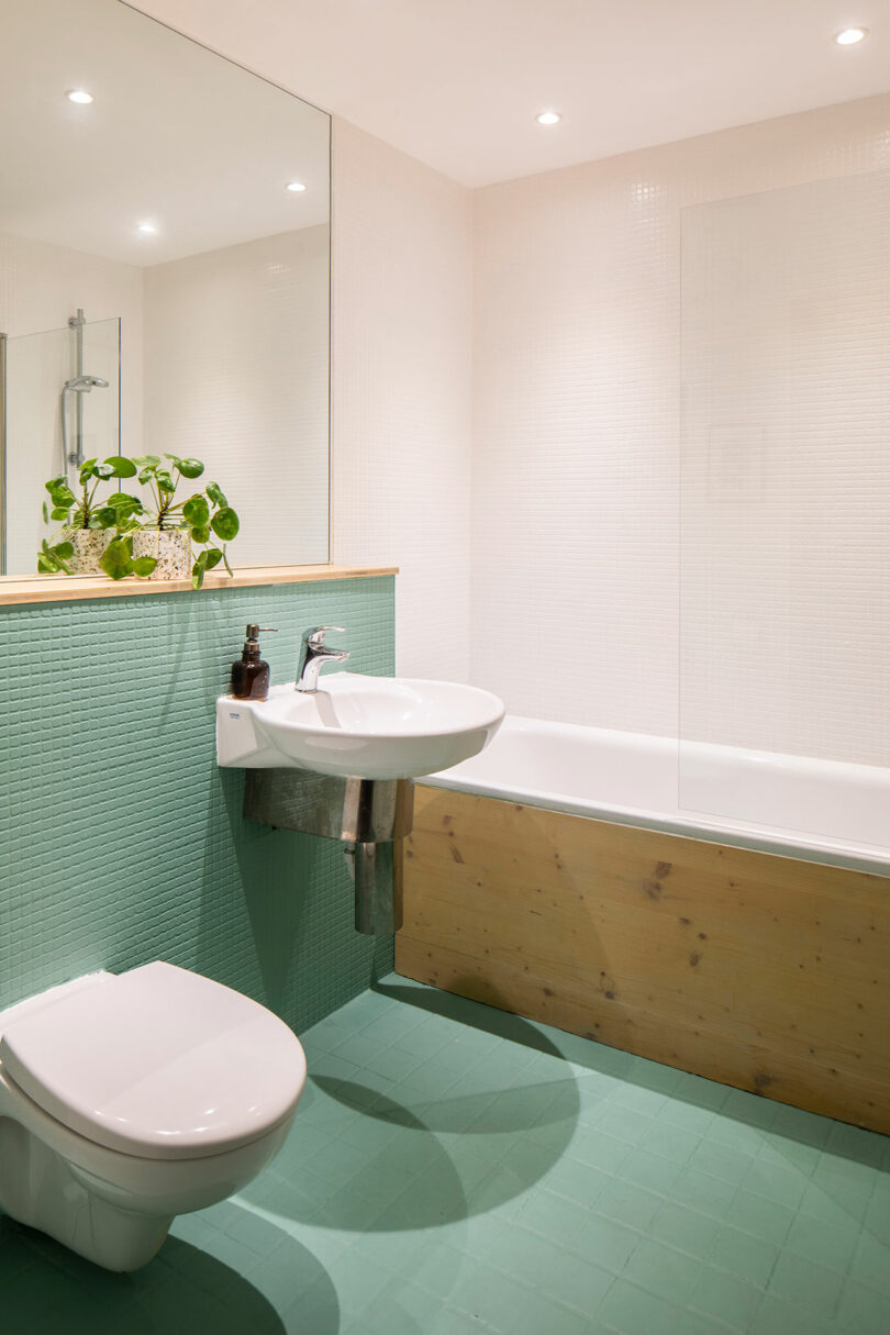 A modern bathroom with a white sink, toilet, and bathtub. The walls and floor are tiled in green, with a plant and soap dispenser on the sink counter. There is a large mirror above the sink.